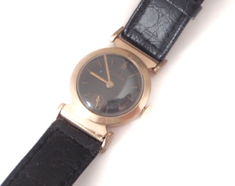 May include: A gold-tone wristwatch with a black face and black leather band. The watch has a round face and a simple design.