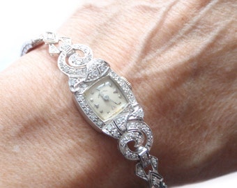 May include: A vintage diamond-encrusted wristwatch with a white face and silver hands. The watch is on a bracelet with a decorative design.