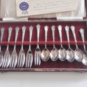 May include: A set of 12 silver-plated teaspoons and forks in a red velvet-lined box. The box has a label that reads "Made and Guaranteed by the Manufacturers Roll and Gold Ltd of Melbourne Australia whose reputation for Quality has been maintained for many years."
