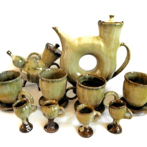 May include: A ceramic tea set in shades of green and brown. The set includes a teapot with a unique circular design, cups, saucers, and small pedestal cups. The pottery has a rustic, handmade appearance.