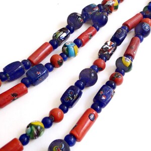 May include: Close-up of two strands of colorful glass bead necklaces. The beads are a mix of blue, red, and multicolored, with various shapes including round, cylindrical, and rectangular. The necklaces are displayed against a white background.