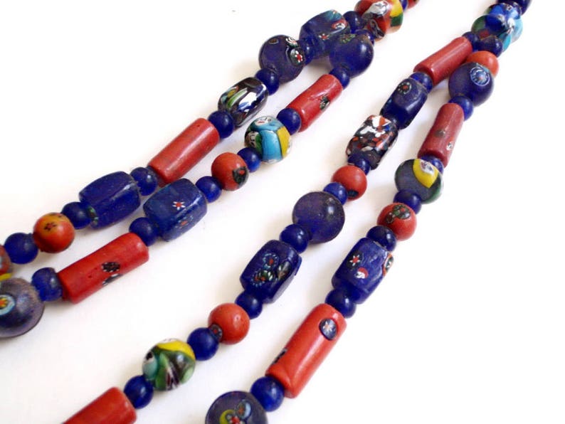 May include: Close-up of two strands of colorful glass bead necklaces. The beads are a mix of blue, red, and multicolored, with various shapes including round, cylindrical, and rectangular. The necklaces are displayed against a white background.