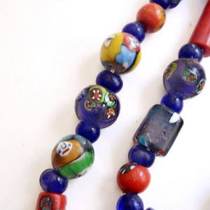 May include: Close-up of a colorful beaded necklace. The necklace features a variety of beads, including round, cylindrical, and rectangular shapes. The beads are in shades of blue, red, yellow, and green, with intricate patterns and designs.
