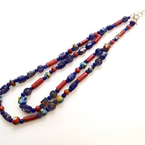 May include: A layered necklace featuring a mix of blue, red, and multicolored beads. The necklace has a gold-colored clasp and is composed of various shapes and sizes of beads, creating a unique and eye-catching design.