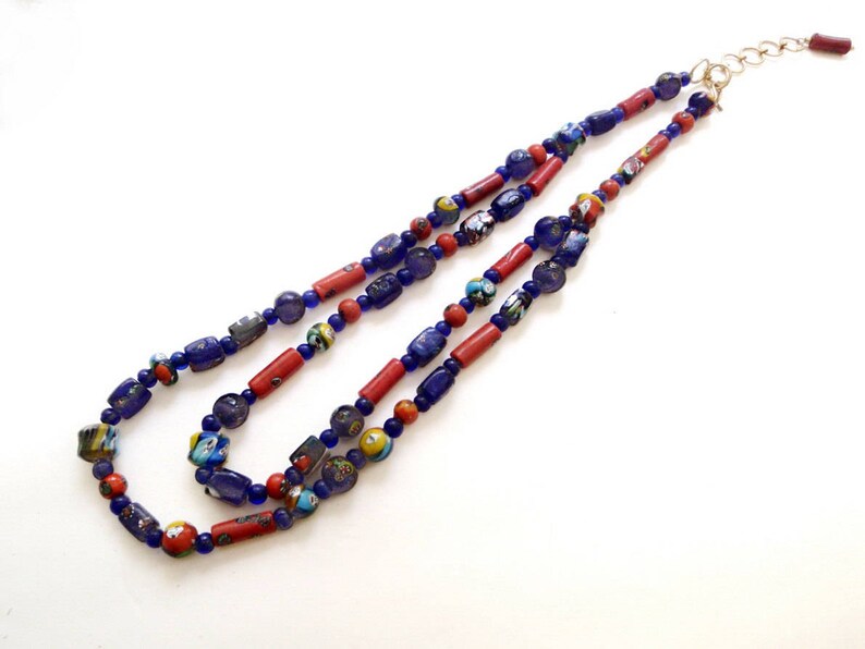 May include: A layered necklace featuring a mix of blue, red, and multicolored beads. The necklace has a gold-colored clasp and is composed of various shapes and sizes of beads, creating a unique and eye-catching design.