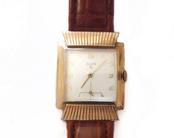 May include: A gold-toned Elgin wristwatch with a white face and black hands. The watch has a square face with a decorative gold bezel and a brown leather band.
