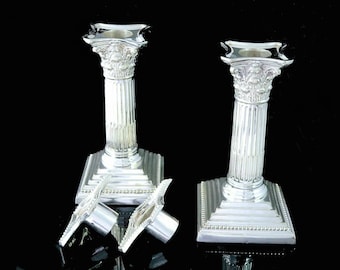 May include: Two silver-colored candle holders with a classical column design. Each holder has a fluted column, a detailed capital, and a stepped base. Two additional silver pieces are in front of the candle holders. The items are on a black background.