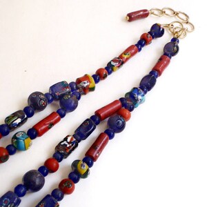 May include: A multi-strand necklace featuring a variety of beads. The necklace includes red, blue, and multicolored glass beads, with a gold-colored chain and clasp. The beads are arranged in a repeating pattern.