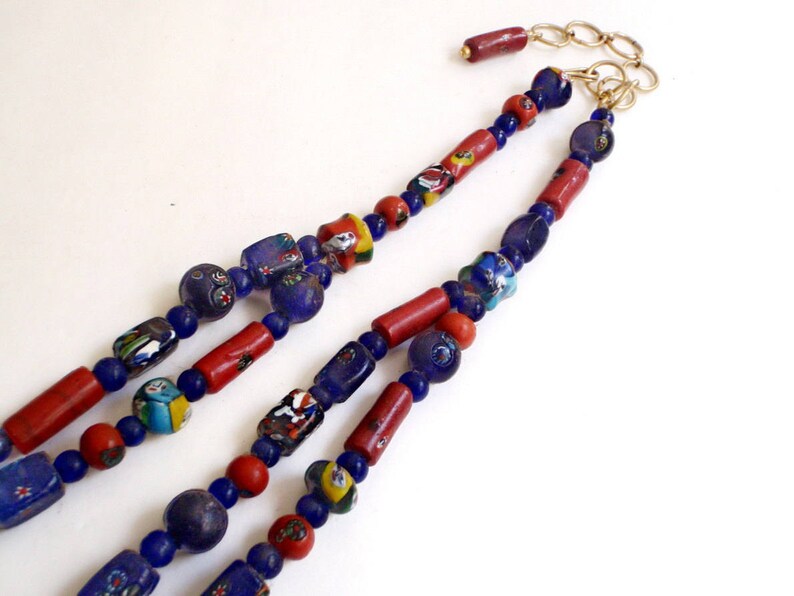 May include: A multi-strand necklace featuring a variety of beads. The necklace includes red, blue, and multicolored glass beads, with a gold-colored chain and clasp. The beads are arranged in a repeating pattern.
