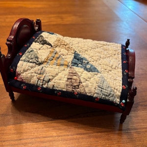 May include: A miniature wooden bed frame with a dark brown finish and four turned posts. The bed is dressed with a quilted coverlet featuring a patchwork pattern of blue, white, and tan fabric, bordered by a dark blue trim with a small red floral print.