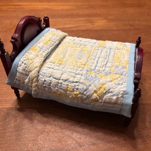 May include: A miniature dollhouse bed with a dark brown wooden frame featuring a curved headboard and turned posts. The bed is dressed with a light blue bed skirt and a quilted coverlet patterned with a yellow and white floral design.