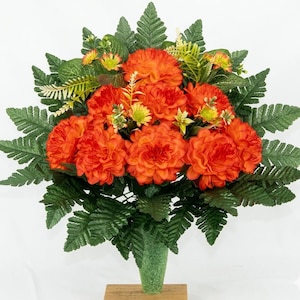 May include: A floral arrangement featuring vibrant orange carnation-like flowers interspersed with green fern fronds and small yellow-tipped accents. The bouquet is gathered into a textured green conical base that sits atop a small wooden block.