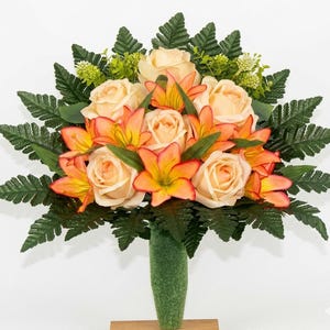 May include: A floral arrangement featuring peach roses and orange lilies, accented by green fern leaves and small green buds. The flowers are arranged in a green vase, creating a vibrant and colorful display.