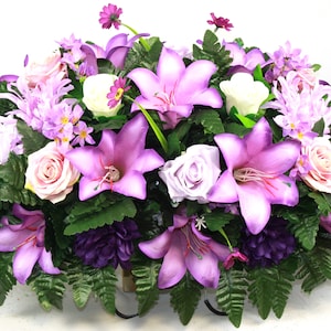 May include: A floral arrangement featuring a variety of artificial flowers in shades of purple, pink, and white. The arrangement includes lilies, roses, and other blossoms, with green foliage. The flowers are arranged in a compact, oval shape.