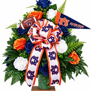 May include: A floral arrangement featuring blue, orange, and white roses, green foliage, and a miniature football with an Auburn logo. A large white ribbon with orange edges and blue Auburn logos cascades down, alongside a blue pennant reading AUBURN TIGERS.