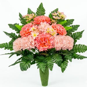 May include: A vibrant floral arrangement featuring coral and light pink dahlias, accented by small yellow flowers and lush green fern leaves. The bouquet is set in a green, textured vase, creating a natural and colorful display.