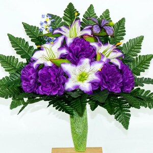 May include: A vibrant floral arrangement featuring purple carnations, white and purple lilies, and green fern leaves. The flowers are arranged in a green glitter vase, creating a colorful display. The arrangement is set against a white background.