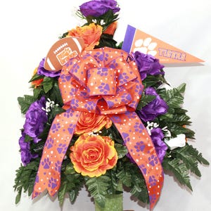 May include: A purple and orange floral arrangement with a football, a bow, and a pennant that says "TIGERS".