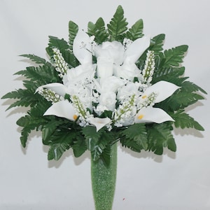 May include: A white artificial flower arrangement with white roses and calla lilies in a green vase. The arrangement is surrounded by green ferns.