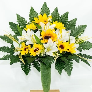 May include: A vibrant floral arrangement featuring sunflowers, white lilies, and green fern leaves. The bouquet is arranged in a green vase, creating a colorful display. The flowers have yellow and white petals with dark brown centers.