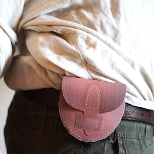 May include: A small, pink leather pouch with a flap closure and a brown leather belt loop. The pouch is worn on a person's hip.