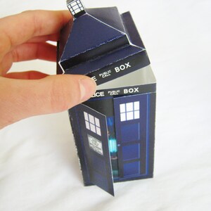May include: A blue and white cardboard model of a police telephone box, also known as a TARDIS, with the words "PUBLIC CALL BOX" printed on the side. The top of the box is open, revealing the interior.