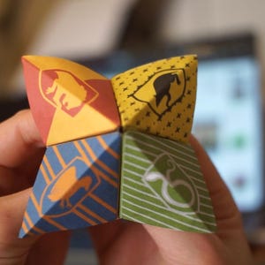 May include: A paper fortune teller with four different colored panels. Each panel has a different image of a creature from the Harry Potter series. The panels are yellow, red, blue, and green.