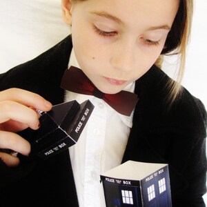 May include: A young person wearing a black blazer and a white shirt with a red bow tie. They are holding a small cardboard model of a blue police box with the text "Police Box" printed on it.