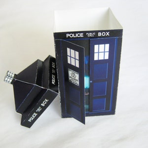 May include: A blue and white cardboard box shaped like a police telephone box. The box has the words "Police Public Call Box" printed on the side. The door is open and the inside of the box is visible.