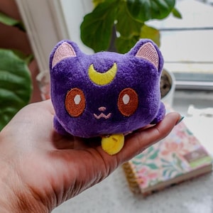 May include: A plush, purple cat-shaped toy with a yellow crescent moon on its forehead and orange eyes. The cat has pink inner ears and a yellow bell. The toy is held in a hand, with a floral notebook visible in the background.