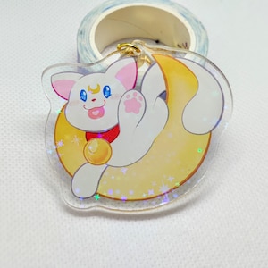 May include: A white cat with pink ears and a blue eye is sitting on a yellow crescent moon with a gold bell around its neck. The background is a white surface with a blue and white tape dispenser.