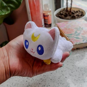 May include: A white plush cat toy with blue eyes, pink ears, and a yellow crescent moon on its forehead. The cat is lying down and has a small yellow collar. The toy is held in a hand.
