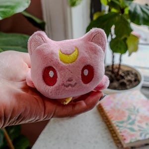 May include: A pink plush cat toy with a yellow crescent moon on its forehead and red eyes. The cat has a small, smiling mouth and is held in a hand. The toy is a cute, soft item, possibly a collectible or a character from a popular series.