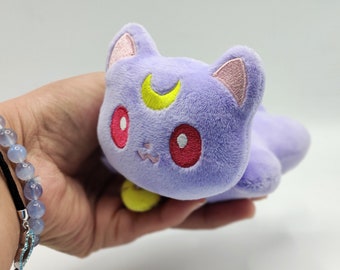 May include: A plush purple cat with pink ears, yellow crescent moon, and red embroidered eyes.