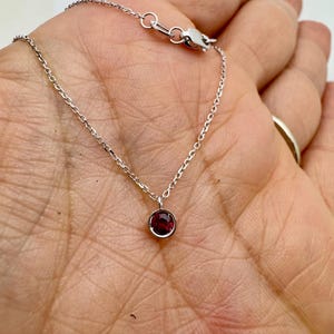 May include: A delicate silver chain necklace with a small, round, dark red gemstone pendant.