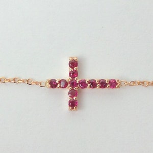 May include: A delicate gold chain bracelet with a small cross charm. The cross is adorned with sparkling red gemstones.