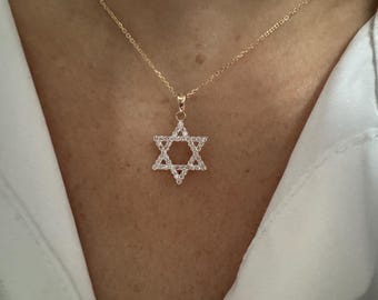 May include: A gold-colored necklace featuring a Star of David pendant. The pendant is encrusted with small, clear gemstones, creating a sparkling effect. The necklace is displayed against a neutral background.