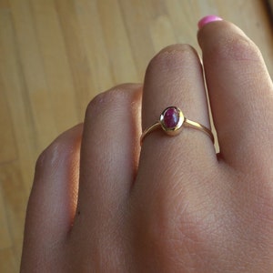 May include: A gold ring with a small, oval-shaped, red gemstone set in a bezel setting.