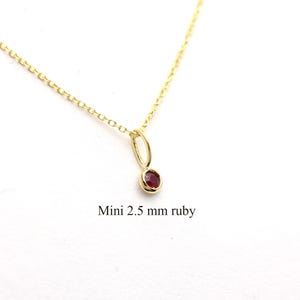 May include: A delicate gold necklace with a small, oval, deep red ruby pendant. The ruby is approximately 2.5 mm in size. The chain is a fine link style. The text "Mini 2.5 mm ruby" is below the pendant.