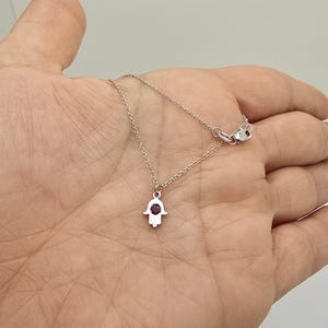 May include: A silver chain necklace with a small silver hamsa hand charm. The hamsa hand has a red gemstone in the centre.