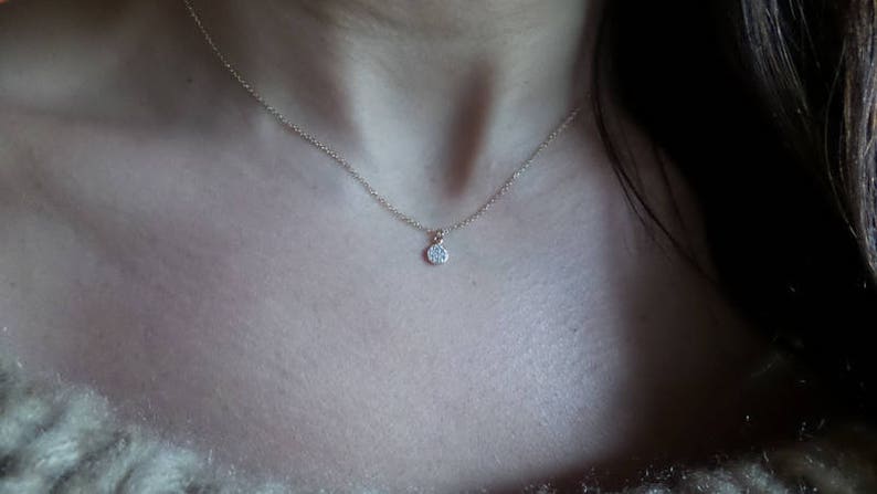 Op de afbeelding: Een delicate zilveren ketting met een klein, rond hangertje met een enkele, heldere edelsteen.