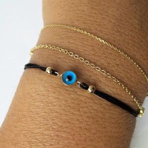 Puede incluir: Un brazalete de cuerda negra con una cuenta dorada y un dije de ojo turco azul. El brazalete está superpuesto con dos brazaletes de cadena dorada.