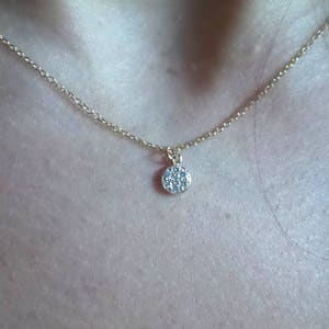 Op de afbeelding: Een delicate gouden ketting met een klein, rond hangertje dat is bezet met kleine, heldere steentjes.