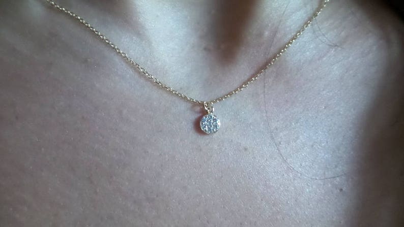 Op de afbeelding: Een delicate gouden ketting met een klein, rond hangertje dat is bezet met kleine, heldere steentjes.
