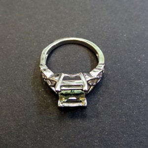 May include: A silver ring with a green gemstone set in a square shape. The ring has a delicate design with intricate details.
