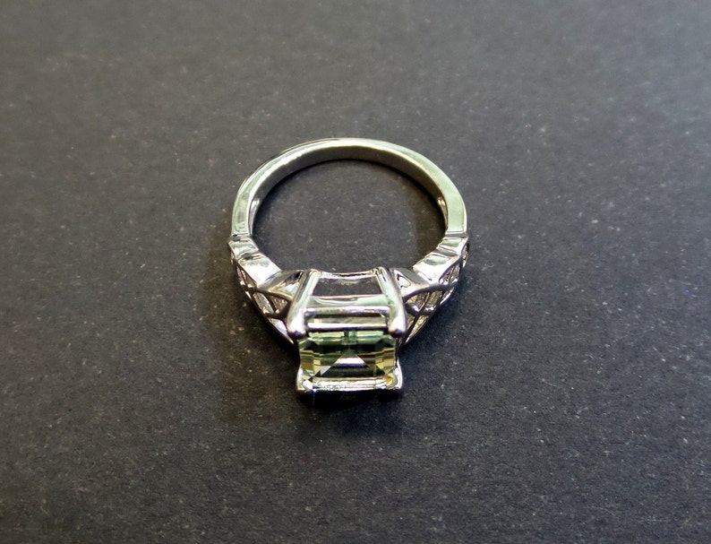 May include: A silver ring with a green gemstone set in a square shape. The ring has a delicate design with intricate details.
