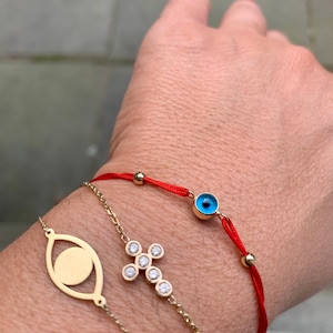 Puede incluir: Tres pulseras en una muñeca. La pulsera superior es de hilo rojo con un dije de ojo turco azul. La pulsera del medio es una cadena de oro con un dije de cruz de diamantes. La pulsera inferior es una cadena de oro con un dije de ojo turco.