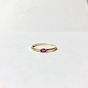 May include: A simple gold band ring with a small, oval-shaped, dark pink gemstone set in the center.