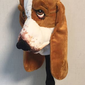 May include: A brown and white plush golf club head cover shaped like a basset hound dog. The head cover has a black nose and brown eyes.