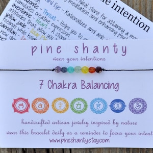 May include: A white card with the text "gemstone intention" and "pine shanty wear your intentions" on it. A black string bracelet with seven colorful beads is on the card. The card also has the text "7 Chakra Balancing" and symbols for each chakra.
